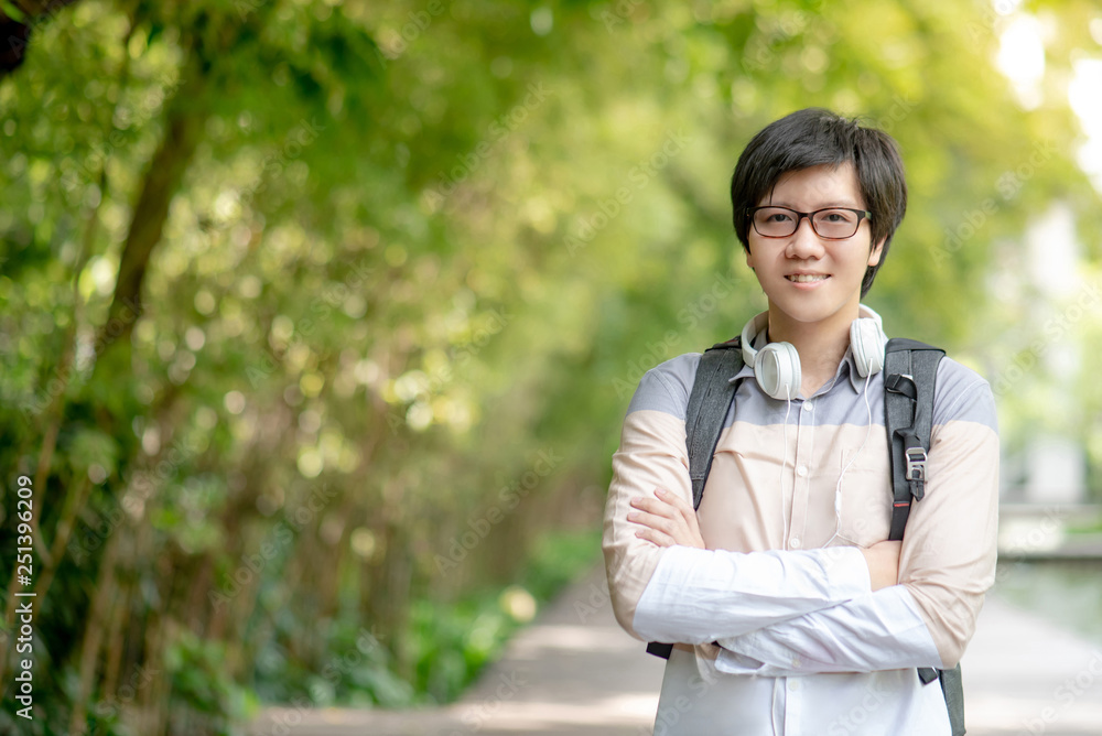Confident Asian man university student with glasses and headphones smiling and crossing arms in the green park of college. Happy life in campus. Education opportunity and scholarship concepts Confident Asian man university student with glasses and headphones smiling and crossing arms in the green park of college. Happy life in campus. Education opportunity and scholarship concepts