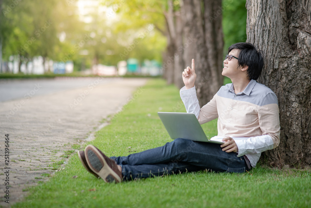 Young Asian man university student with glasses and laptop computer sitting on green grass pointing finger up in the college park. Education opportunity or scholarship. Idea and inspiration concepts Young Asian man university student with glasses and laptop computer sitting on green grass pointing finger up in the college park. Education opportunity or scholarship. Idea and inspiration concepts