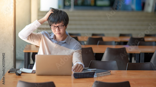 Young Asian man university student with glasses feeling frustrated while using laptop computer in college library. Campus lifestyle in education building. Research problem and solution concepts