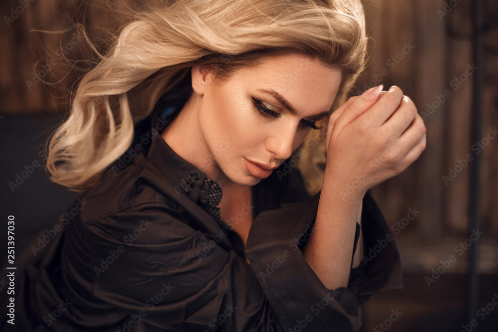 Fototapeta premium Gorgeous model. Blond woman portrait in black shirt. Fashionable girl with beauty makeup and curly hair style posing in wooden dark interior.