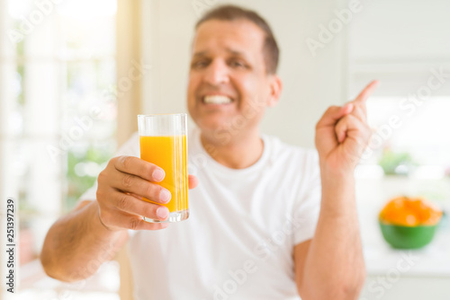 Middle age man drinking a glass of orange juice at home very happy pointing with hand and finger to the side