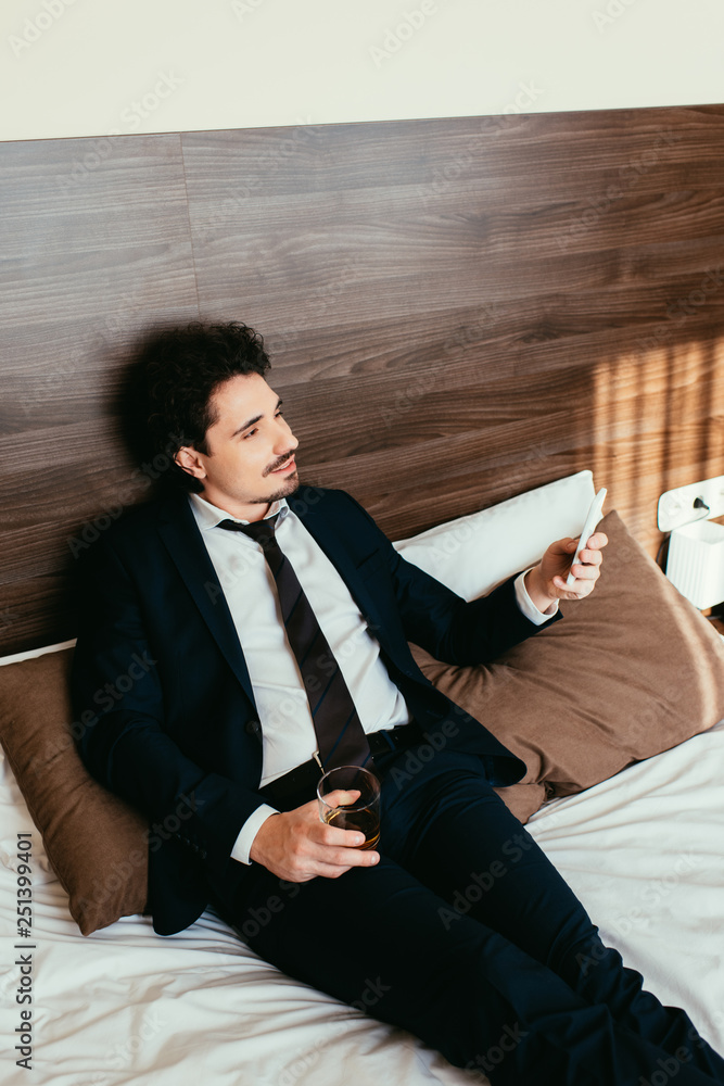 businessman using smartphone and holding glass of cognac on bed in hotel room