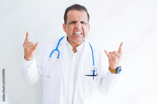 Middle age doctor man wearing stethoscope and medical coat over white background shouting with crazy expression doing rock symbol with hands up. Music star. Heavy concept. © Krakenimages.com