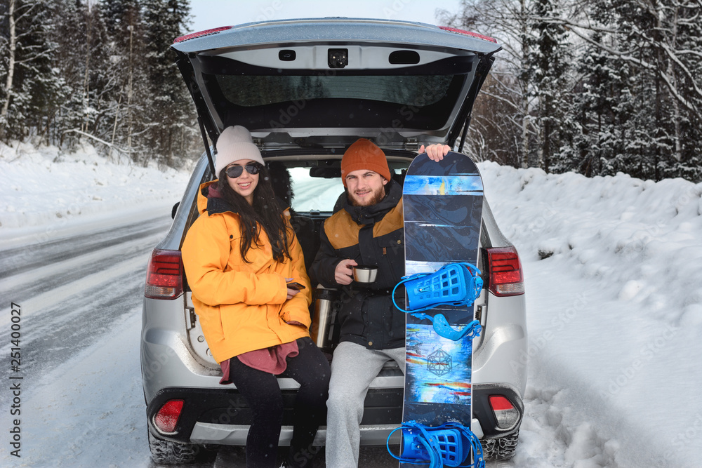 Young couple is sitting in the back trunk of suv car on winter road and drinking hot tea from thermos. Return from mountain ski resort. Man holding his snowboard. travelling lifestyle concept.