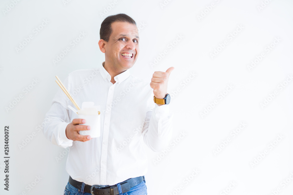 Middle age man eating asian noodles from take away carton pointing and showing with thumb up to the side with happy face smiling Middle age man eating asian noodles from take away carton pointing and showing with thumb up to the side with happy face smiling