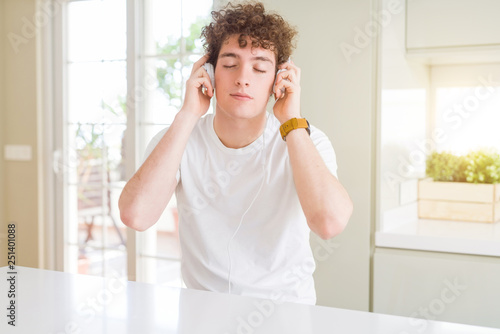 Young man listening to music wearing headphones at homes with serious expression on face. Simple and natural looking at the camera.