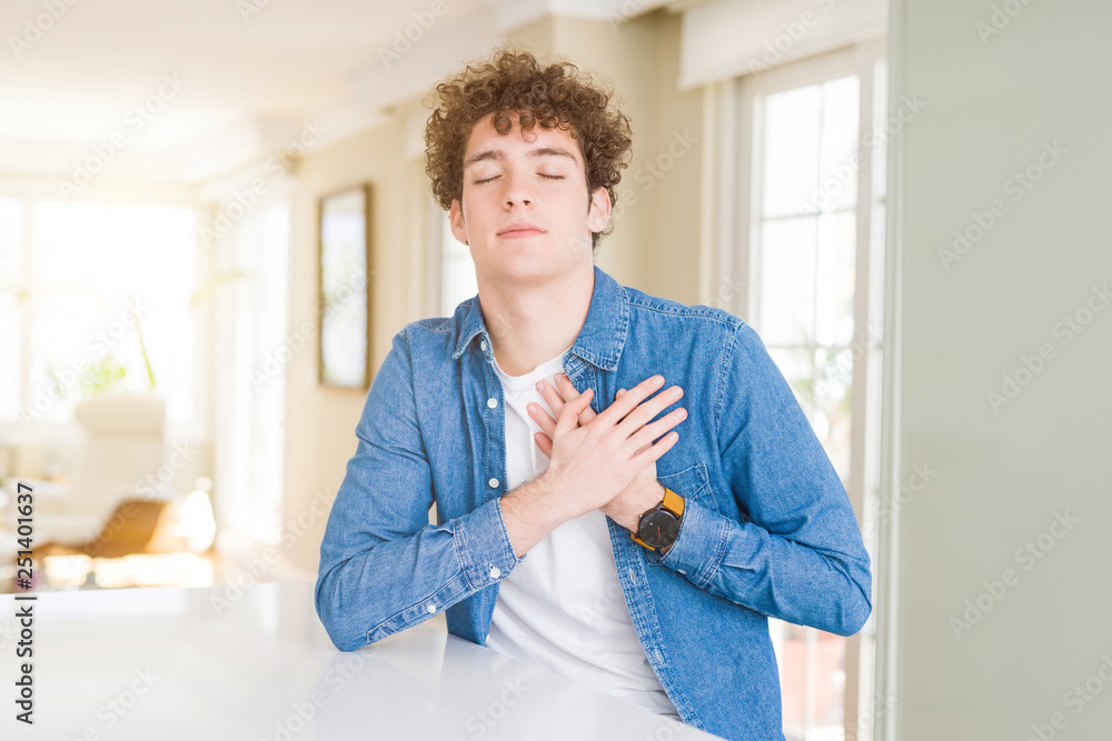 Young handsome man wearing casual denim jacket at home smiling with hands on chest with closed eyes and grateful gesture on face. Health concept.