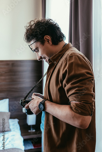 smiling photographer in eyeglasses looking at photo camera in hotel room