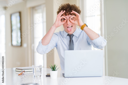 Young business man working with computer laptop at the office doing ok gesture like binoculars sticking tongue out, eyes looking through fingers. Crazy expression.