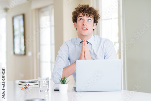 Young business man working with computer laptop at the office begging and praying with hands together with hope expression on face very emotional and worried. Asking for forgiveness. Religion concept.