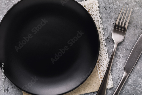Empty ceramic plate black color with a fork and a knife on a table, top view. Food background