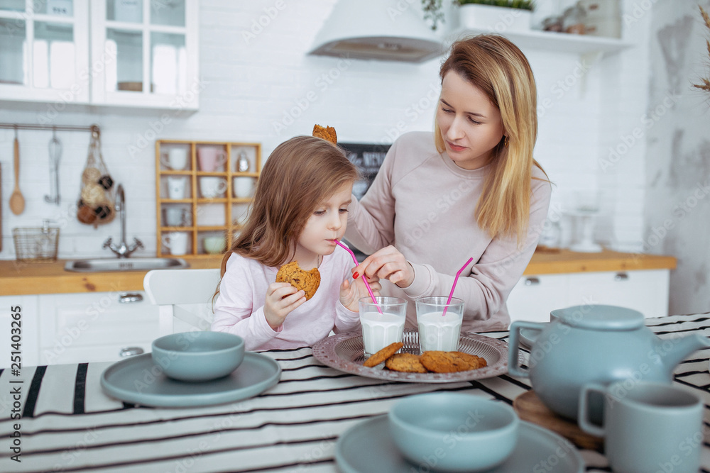 Happy little girl and her beautiful young mother have breakfast together in a white kitchen. They drink milk and eat cookies. Maternal care and love. Horizontal photo Happy little girl and her beautiful young mother have breakfast together in a white kitchen. They drink milk and eat cookies. Maternal care and love. Horizontal photo