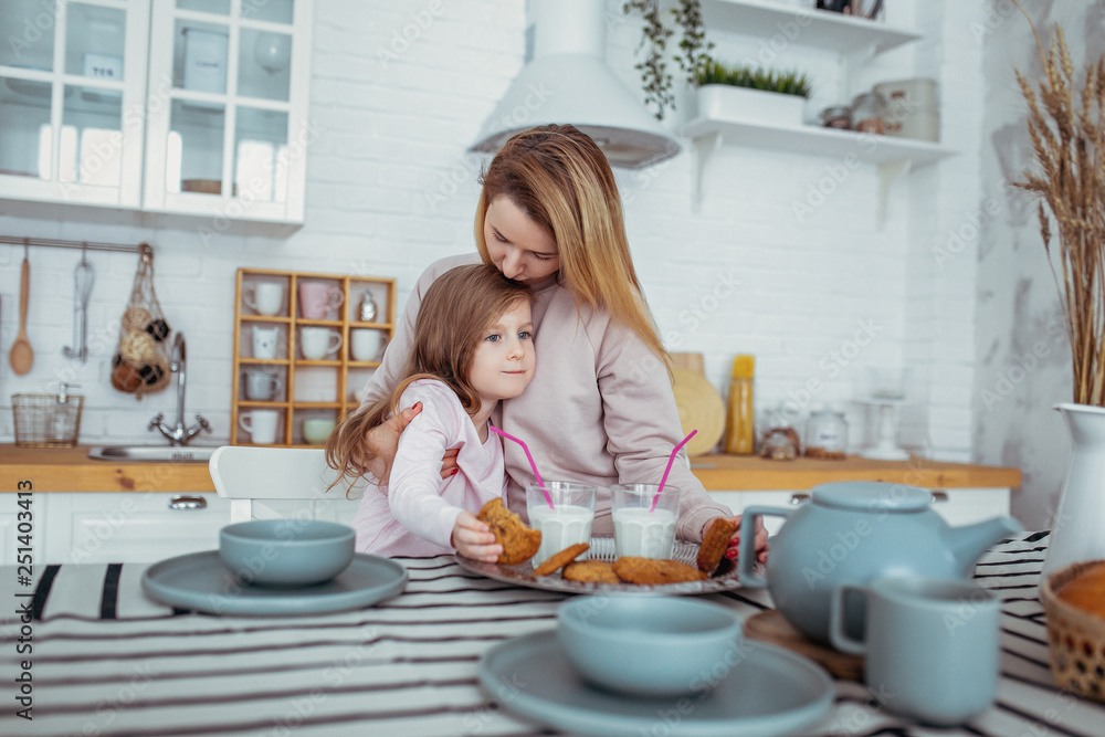 Happy little girl and her beautiful young mother have breakfast together in a white kitchen. Mom hugs and kisses daughter. Maternal care and love. Horizontal photo