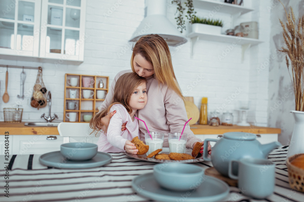 Happy little girl and her beautiful young mother have breakfast together in a white kitchen. Mom hugs and kisses daughter. Maternal care and love. Horizontal photo Happy little girl and her beautiful young mother have breakfast together in a white kitchen. Mom hugs and kisses daughter. Maternal care and love. Horizontal photo