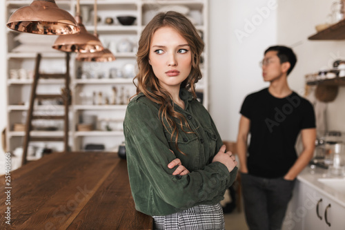 Young beautiful woman with wavy hair holding hands together sadly looking aside with asian man on background. International couple quarrel spending time on kitchen