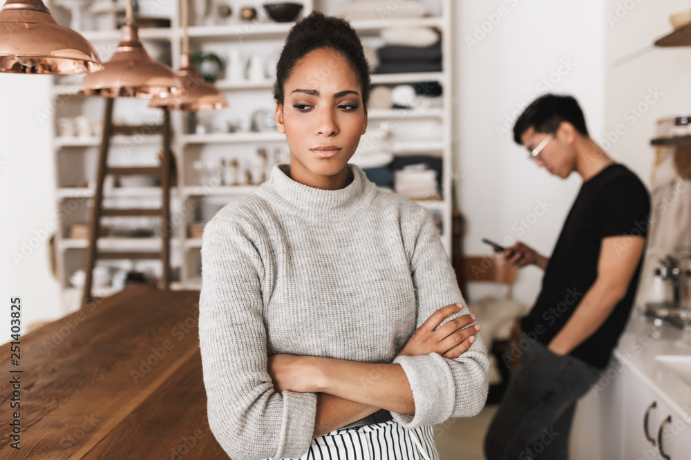 Upset african american woman holding hands together sadly looking aside with asian man holding cellphone on background. Young international couple in quarrel spending time on kitchen Upset african american woman holding hands together sadly looking aside with asian man holding cellphone on background. Young international couple in quarrel spending time on kitchen
