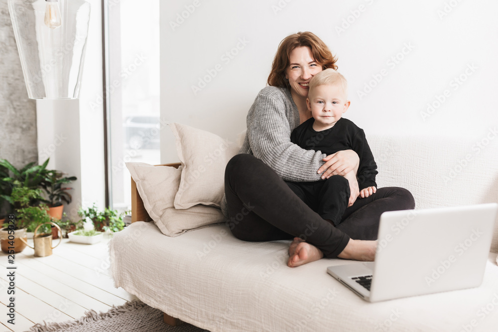 Young smiling woman sitting on sofa with her little beautiful son happily looking in camera. Mom and baby boy joyfully spending time with laptop in living room at home Young smiling woman sitting on sofa with her little beautiful son happily looking in camera. Mom and baby boy joyfully spending time with laptop in living room at home