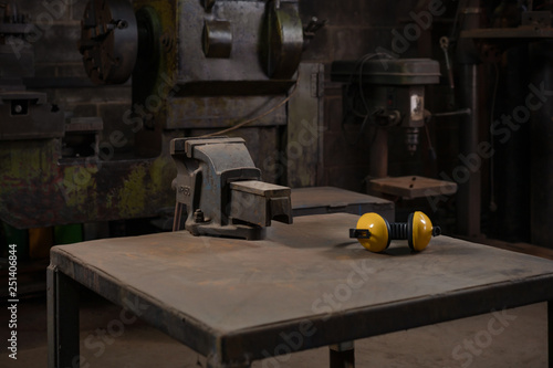 Industrial mechanical cast iron vice bolted to a metal work station table in a dark traditional old style repair workshop next to yellow ear defenders