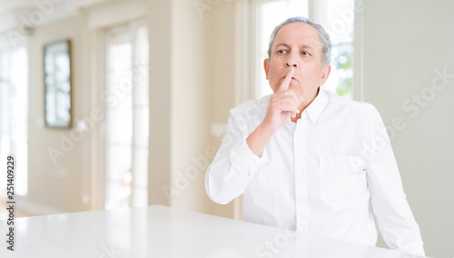 Handsome senior man at home asking to be quiet with finger on lips. Silence and secret concept.