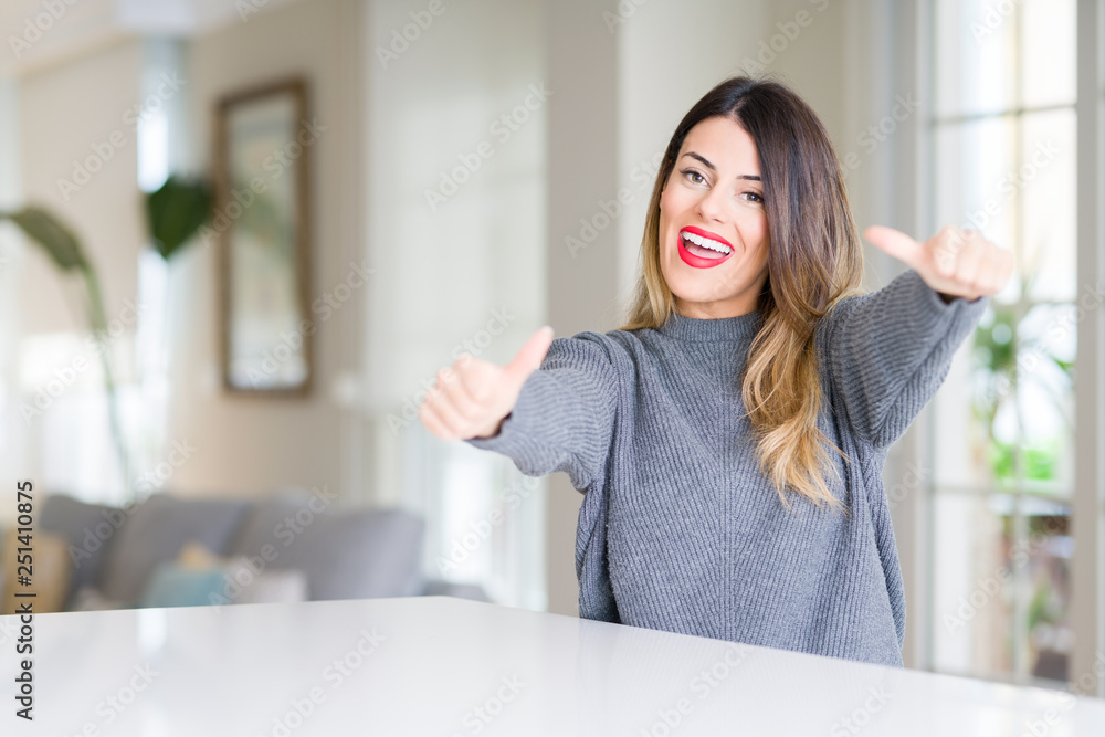 Young beautiful woman wearing winter sweater at home approving doing positive gesture with hand, thumbs up smiling and happy for success. Looking at the camera, winner gesture.