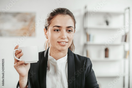 Portrait of a brunette holds white cup of tea in hand. She looks down. Girl smiles a bit photo