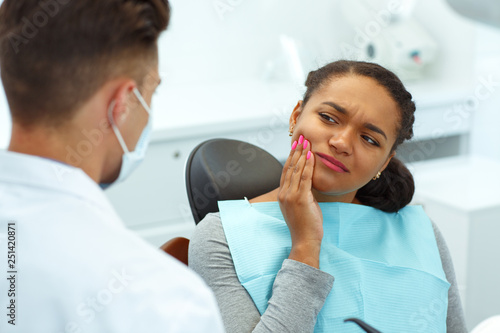 Young woman having toothache, covering mouth by hand and shrugged from pain, sitting in dental chair. Professional dentist wearing in mask and protective glasses, helping patient to cure.