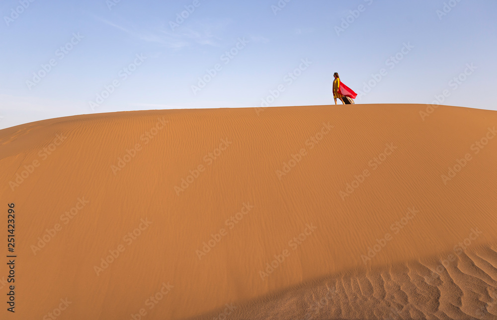 A girl in a yellow dress with a red scarf walks through the desert with a suitcase in her hands. Summer holidays, wild beach, yellow sand, a trip to the unexplored sandy desert