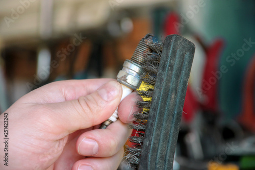 Cleaning a car engine spark plug with wire brush in car garage. Mechanic is brushing used spark plug with a stainless steel brush in auto mechanics workshop. Worker with service tools in hand.