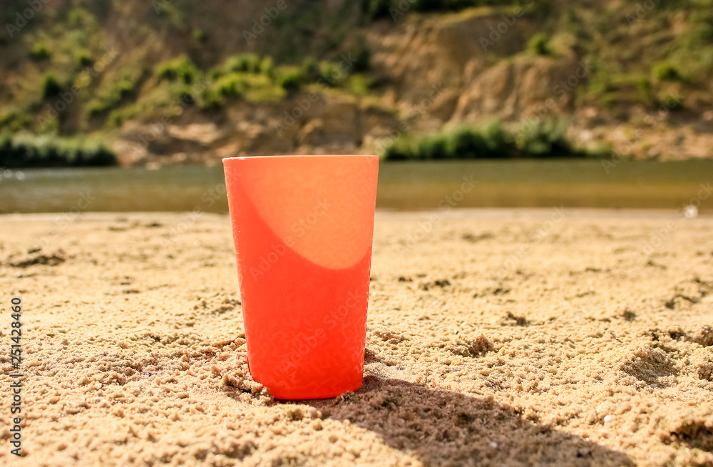 an orange glass stands in the sand, on the shore of the pond an orange glass stands in the sand, on the shore of the pond