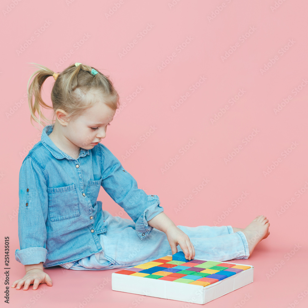 A charming little girl in denim clothes on a pink background puts wooden multi-colored cubes in a white box. Copy space. A charming little girl in denim clothes on a pink background puts wooden multi-colored cubes in a white box. Copy space.