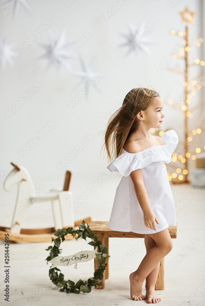 Obraz premium A beautiful little girl sitting on a wooden stool in a beautiful dress in the white room