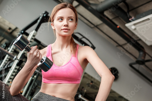 Young girl in gym healthy lifestyle sitting on bench with dumbbell looking aside curious close-up