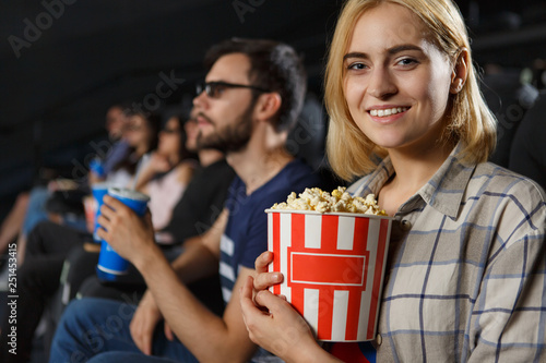 Positive smiling girl in checkered shirt looking at camera and posing in cinema hall. Young lady eating popcorn and watching new interesting movie on weekends. Concept of entertainment.
