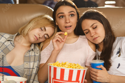Female friends on film watching at cinema theater.  Shocked girl watching horror movie and expecting final while eating popcorn. Two other girls sleeping near during boring film. Concept of  leisure.