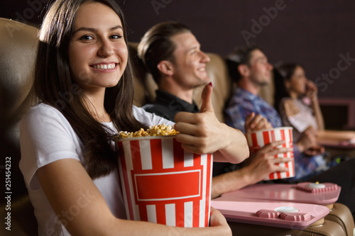 Happy brunette sitting in cinema hall, eating popcorn and showing sign okay while watching interesting film. Pretty girl in white shirt enjoying funny movie on weekends. Concept of leisure and fun.