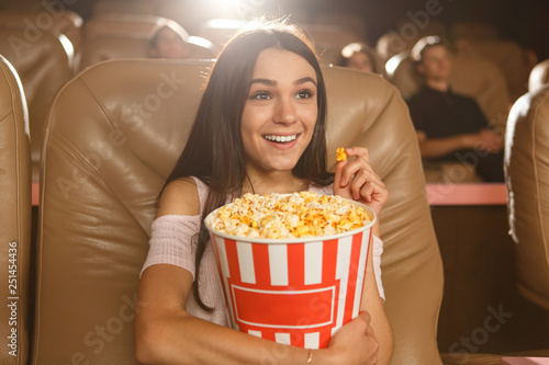 Positive and beautiful dark haired girl enjoying watching interesting film at cinema. Pretty female viewer in white shirt and jeans looking at screen  keeping popcorn and smiling.
