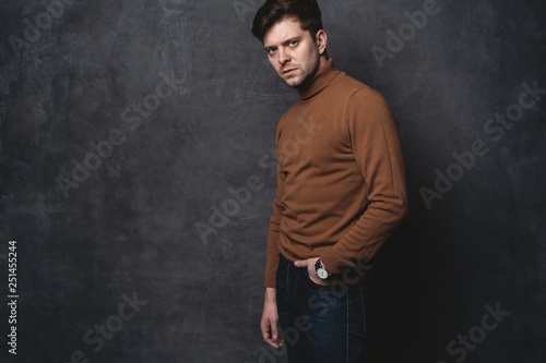Handsome young man on gray background looking at camera. Portrait of young man with hands in pockets leaning against gray wall.