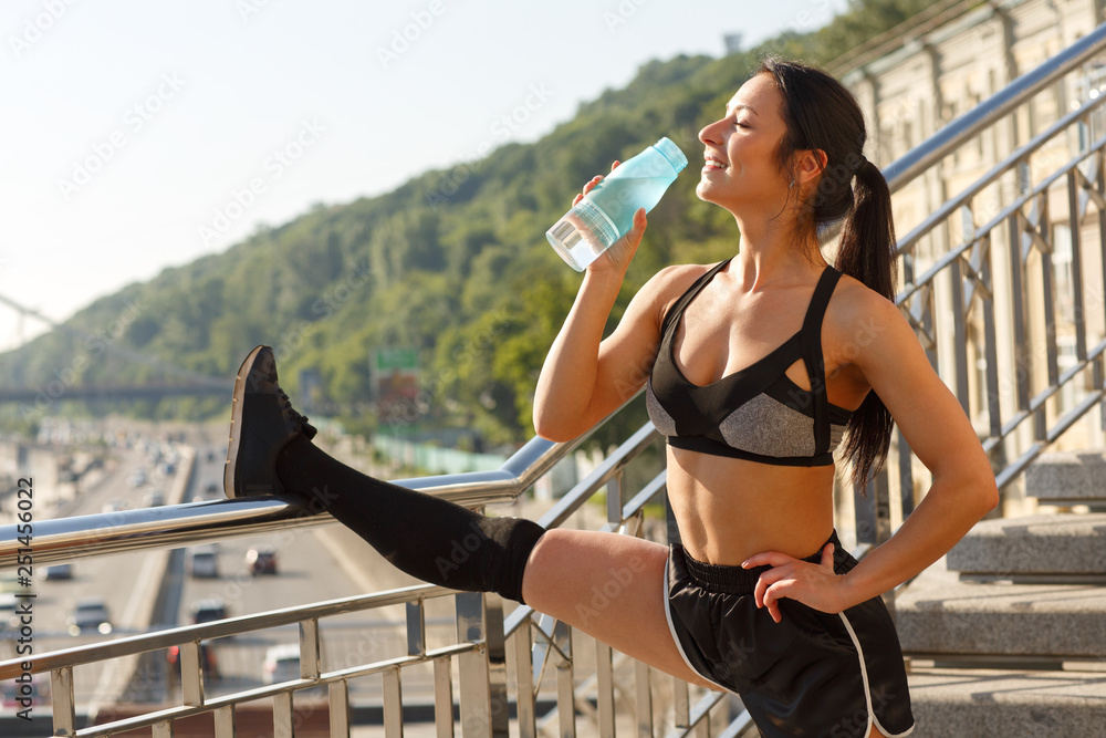 Sporty woman with fit body wearing black tracksuit stretching and drinking  water after hard training outdoor. Female athlete standing on stairs,  keeping leg on railings and relaxing. Stock Photo
