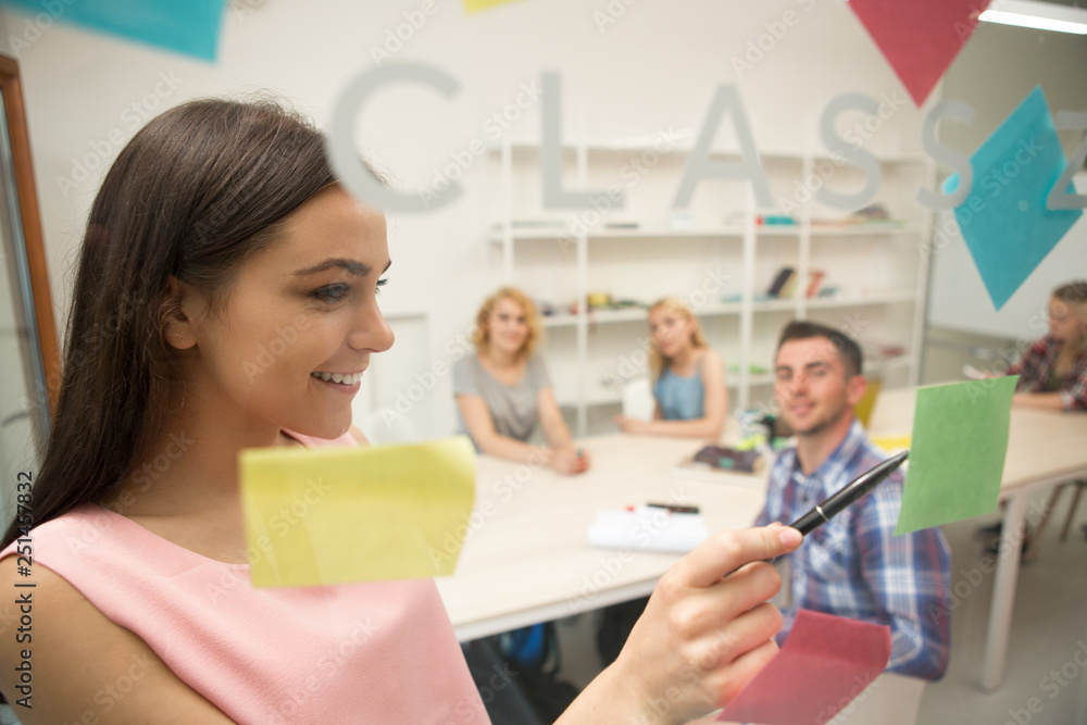 Girl standing and writing on stickers at class glass board at university.