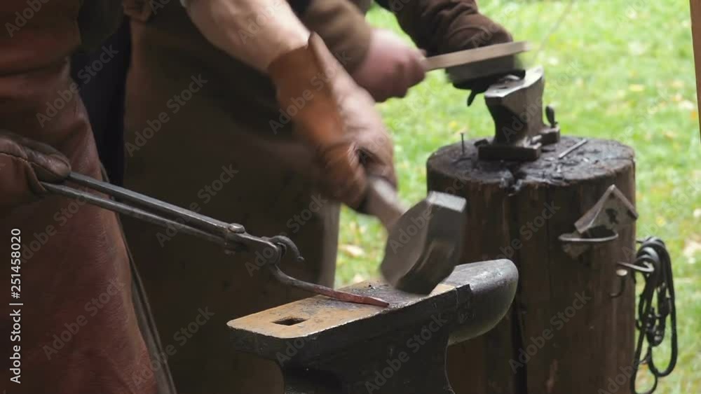 Two blacksmiths in the production process of metal products handmade on the openair workshop. One of blacksmiths strikes with a hammer on metal, another cleans the workpiece from the scale and soot Two blacksmiths in the production process of metal products handmade on the openair workshop. One of blacksmiths strikes with a hammer on metal, another cleans the workpiece from the scale and soot