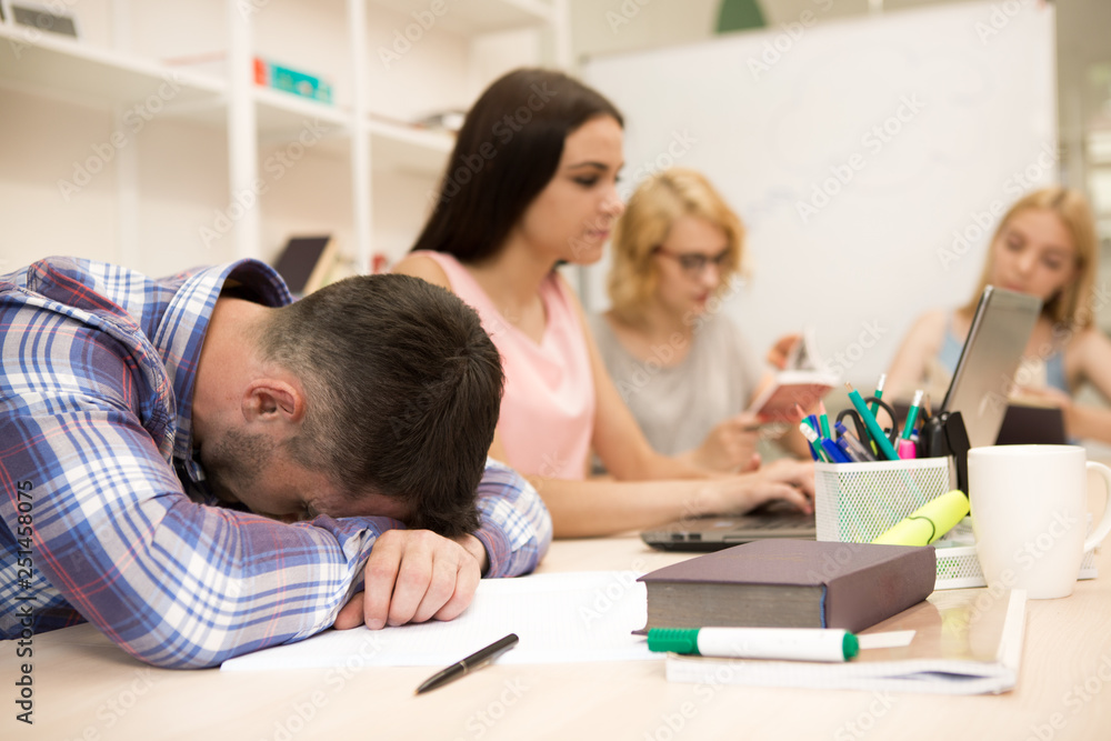 View of male student tired student sleeping on his books, during lecture at university. Group of students sitting at table, reading book on blur background in class. View of male student tired student sleeping on his books, during lecture at university. Group of students sitting at table, reading book on blur background in class.