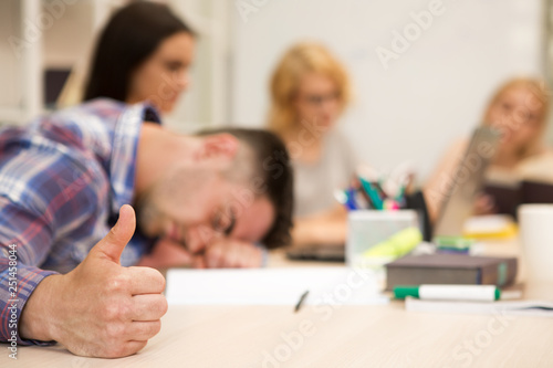 Sleeping boy gesturing thumb up, while group of student, studying at blurred background.