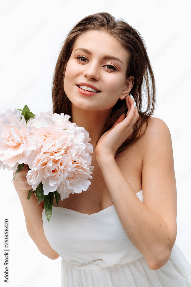 Portrait beautiful young girl smiling and posing with artificial flowers on white background in white dress. Portrait beautiful young girl smiling and posing with artificial flowers on white background in white dress.
