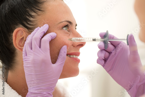 Closeup of hands of beautician making injection in skin around eyes in beauty salon. Hands of doctor in protective gloves doing shot to smiling woman looking away. Concept of lifting and skin care..