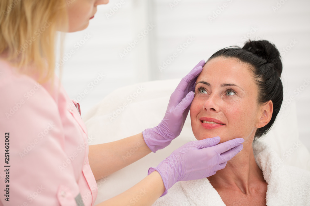 Professional female cosmetologist examining face skin of beautiful woman in beauty parlor. Pretty patient in white robe looking at serious doctor in uniform and protective gloves.
