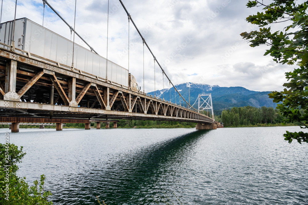 Fototapeta premium Heavy Truck crossing a Suspesion Bridge Spanning a Large River in the Mountains on a Cloudy Summer Day