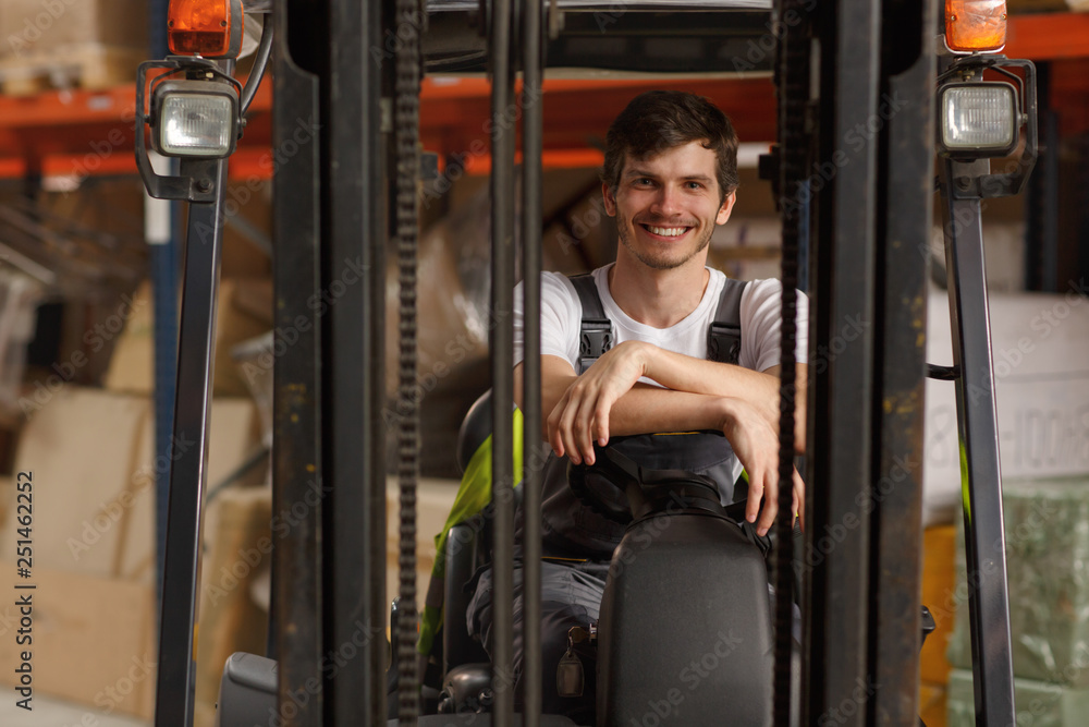 Front view of loader sitting in forklift, posing and smiling. Professional worker wearing uniform and white t shirt. Background of warehouse with many boxes and goods. Front view of loader sitting in forklift, posing and smiling. Professional worker wearing uniform and white t shirt. Background of warehouse with many boxes and goods.