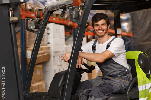 Good looking loader sitting in forklift, posing and smiling. Professional worker wearing uniform and white t shirt. Background of warehouse with many boxes and goods. photo