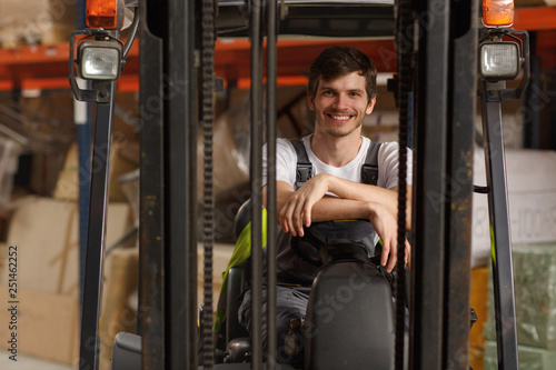 Front view of loader sitting in forklift, posing and smiling. Professional worker wearing uniform and white t shirt. Background of warehouse with many boxes and goods.