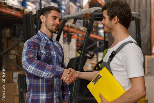 Two men making handshake in warehouse, looking each other. Worker in white t shirt and uniform making deal with customer. Background of forklift. Concept of purchase.